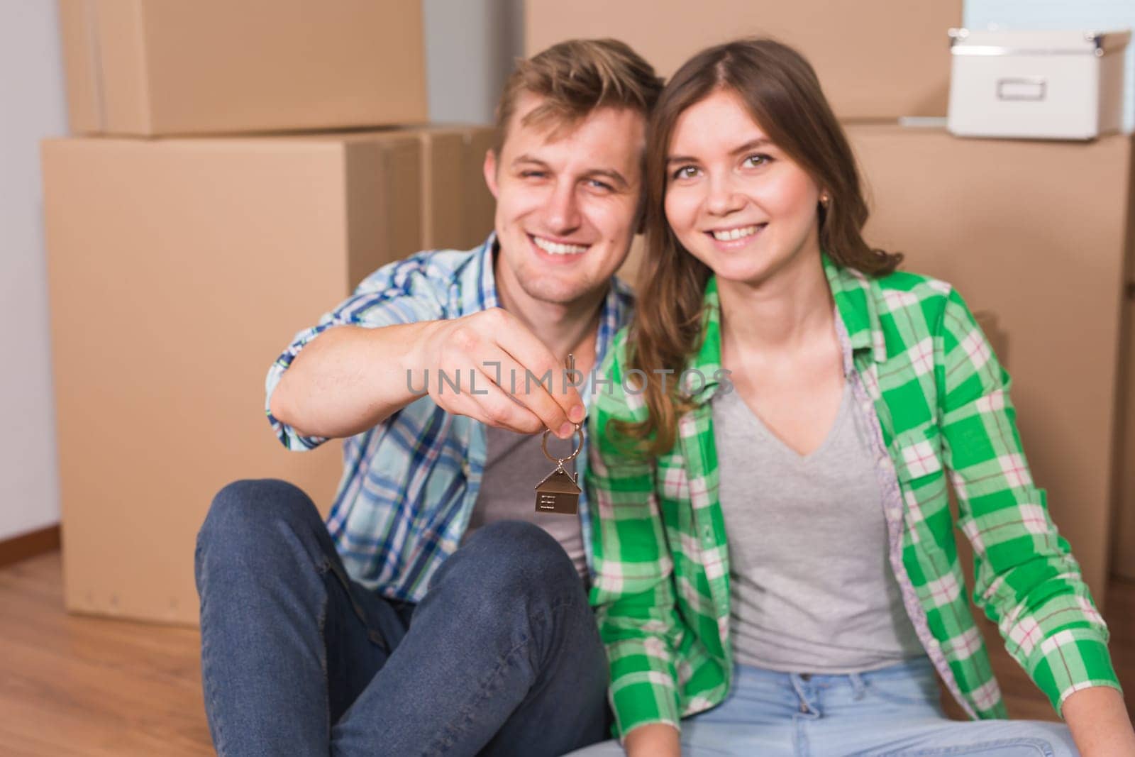 Happy young couple sitting on floor near boxes. Young family moving to new home. Woman and man showing keys and smiling at camera.
