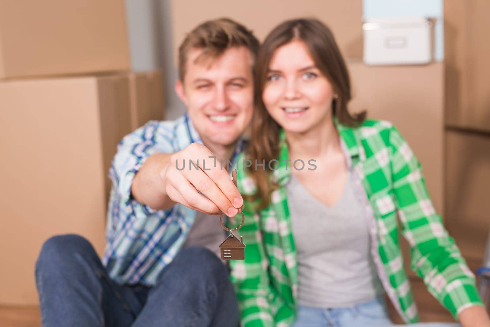 Happy young couple sitting on floor near boxes. Young family moving to new home. Woman and man showing keys and smiling at camera by Satura86