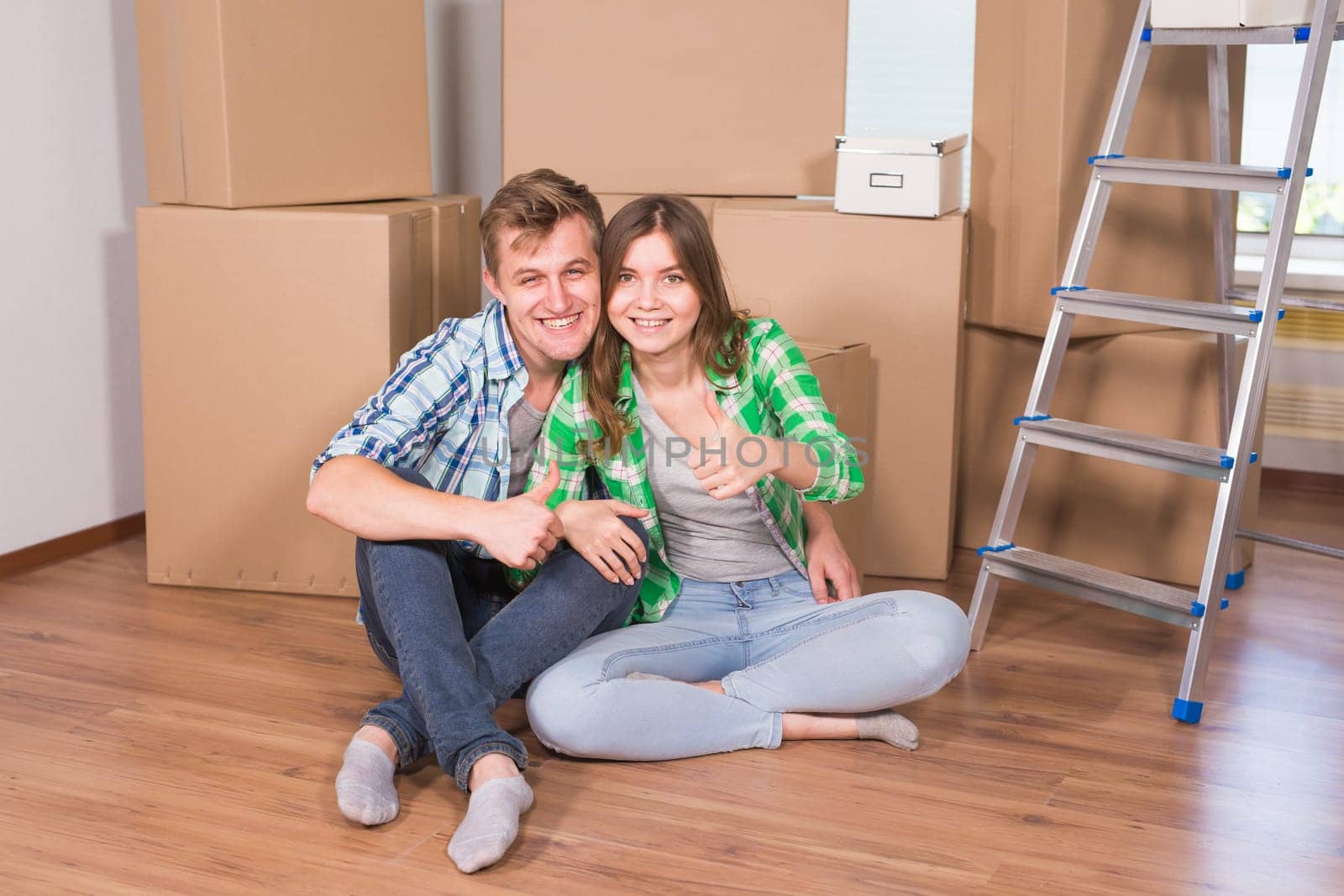 Moving to a new house and repairs in the apartment. Love couple showing a thumbs up and sitting in an empty apartment among boxes.