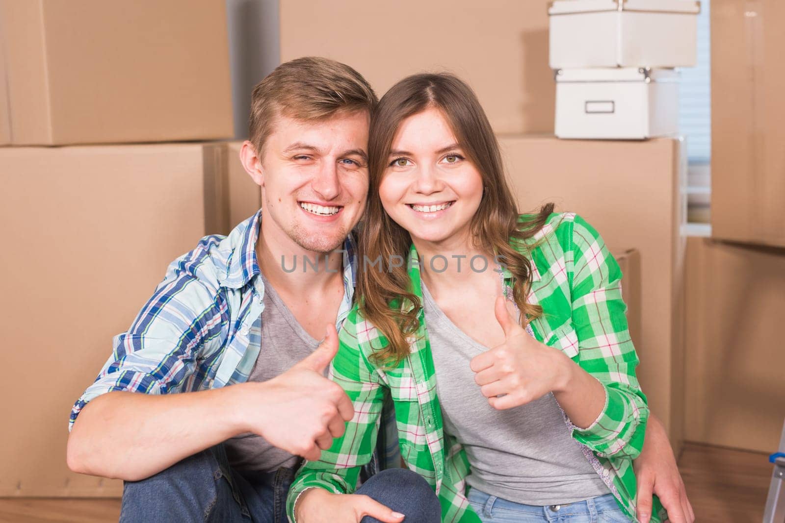 Moving to a new house and repairs in the apartment. Love couple showing a thumbs up and sitting in an empty apartment among boxes by Satura86