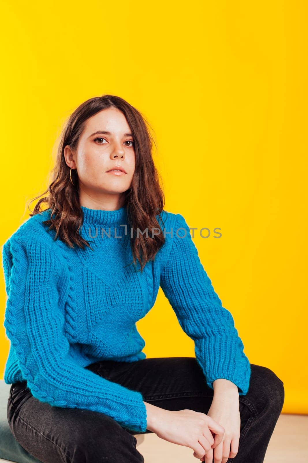 a woman in a warm sweater in winter clothes sits at a yellow background by Simakov