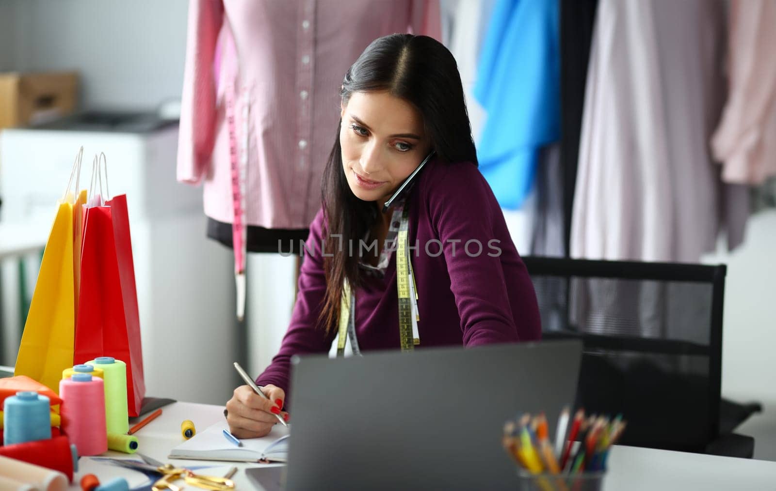Adult caucasian beauty businesswoman takes an order by phone looks at laptop screen and makes notes in notebook an office background. Delivery and tailoring clothes concept.
