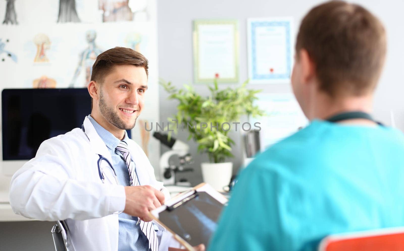 Portrait of smart physician looking at friend with trustful smile. Medical men sitting in big clinic office and discussing treatment. Medicine and healthcare concept
