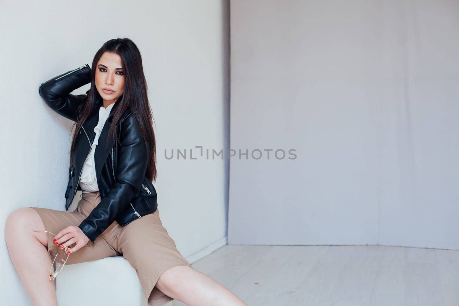 a brunette woman in a leather jacket sits on a low chair by Simakov