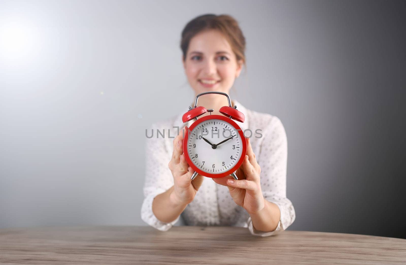 Focus on red alarm clock. Beautiful model in white blouse with curly brunette hair indicating on time. Copy space in left side. Isolated on grey background