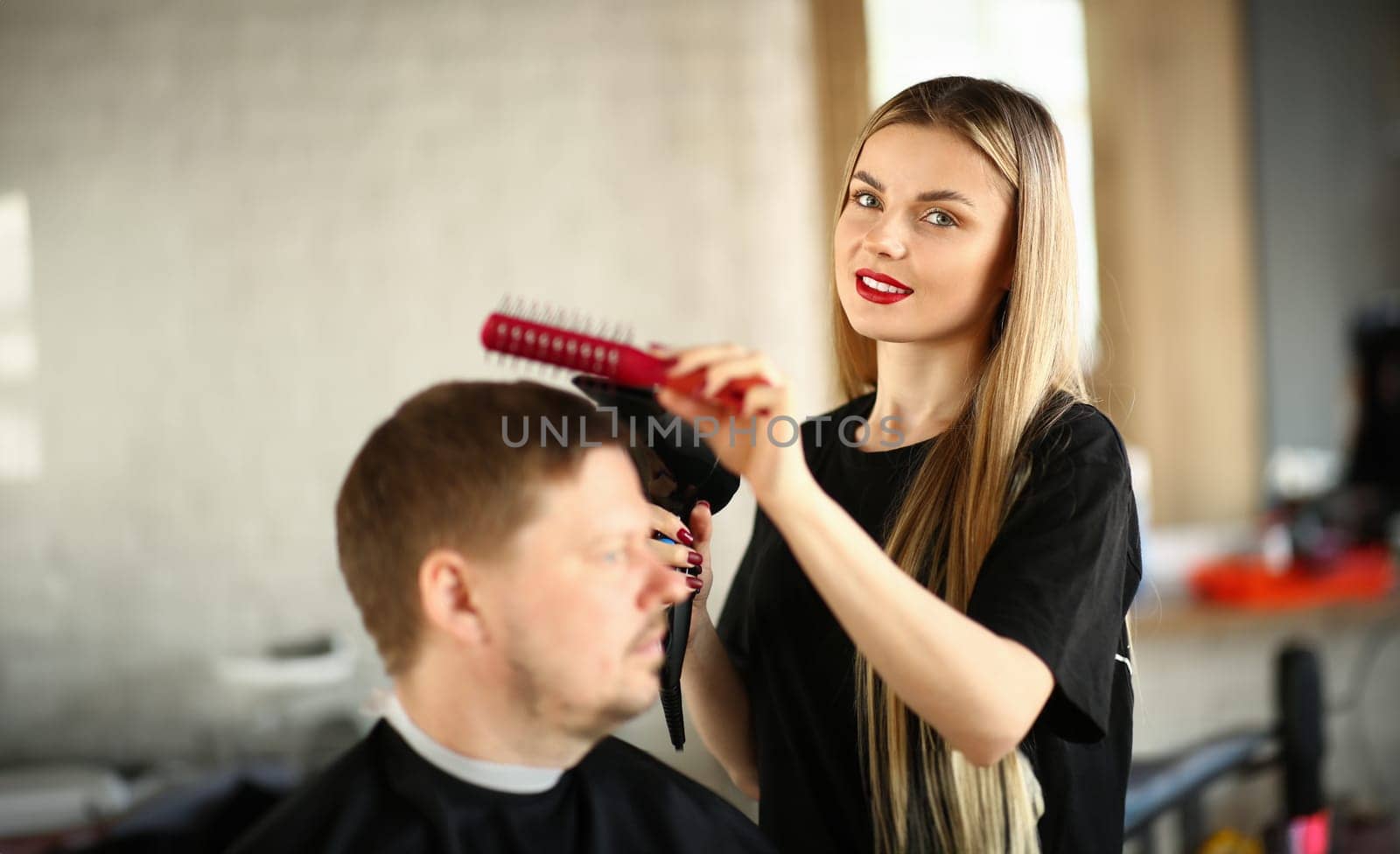 Hairdresser Combing Male Hair and Blowing by Dryer. Young Hairstylist Drying Man Haircut. Woman Beautician Styling Hairdo for Guy Sitting in Beauty Salon. Blonde Stylist Making Hairstyle in Studio