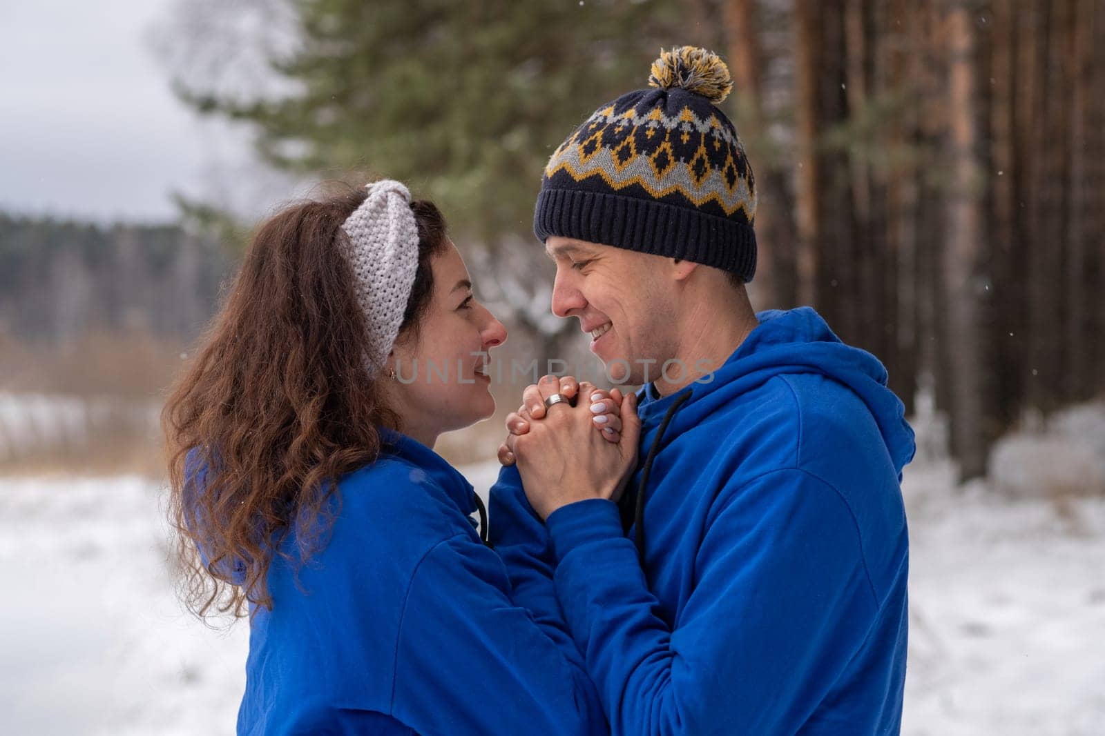 Outdoor happy couple in love posing in cold winter weather. A man and a woman in blue hoodies. Emotional young couple having fun while walking by winter forest, loving man hugging his laughing woman