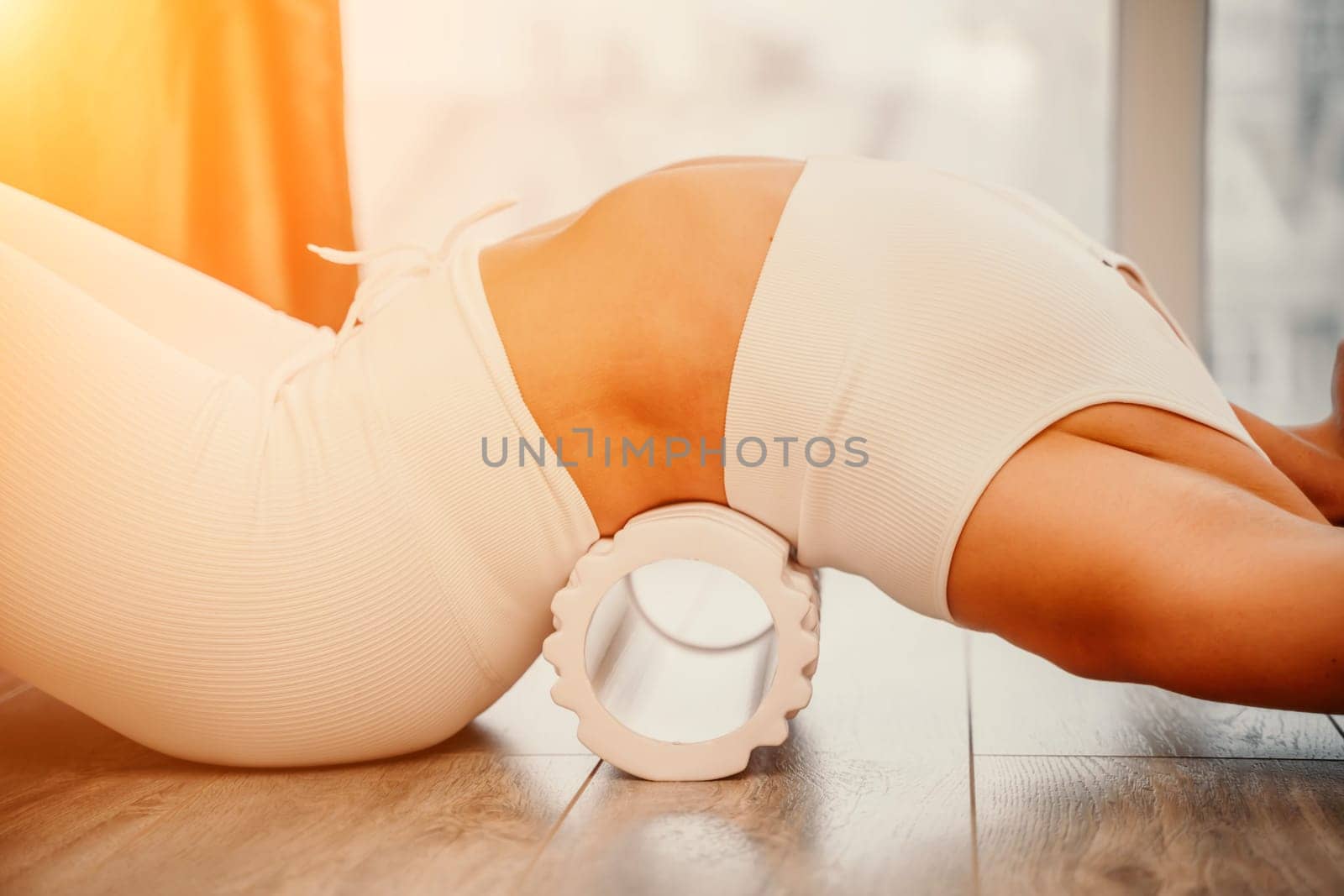 Adult athletic woman, in white bodysuit, performing fascia exercises on the floor - caucasian woman using a massage foam roller - a tool to relieve tension in the back and relieve muscle pain - the concept of physiotherapy and stretching training.