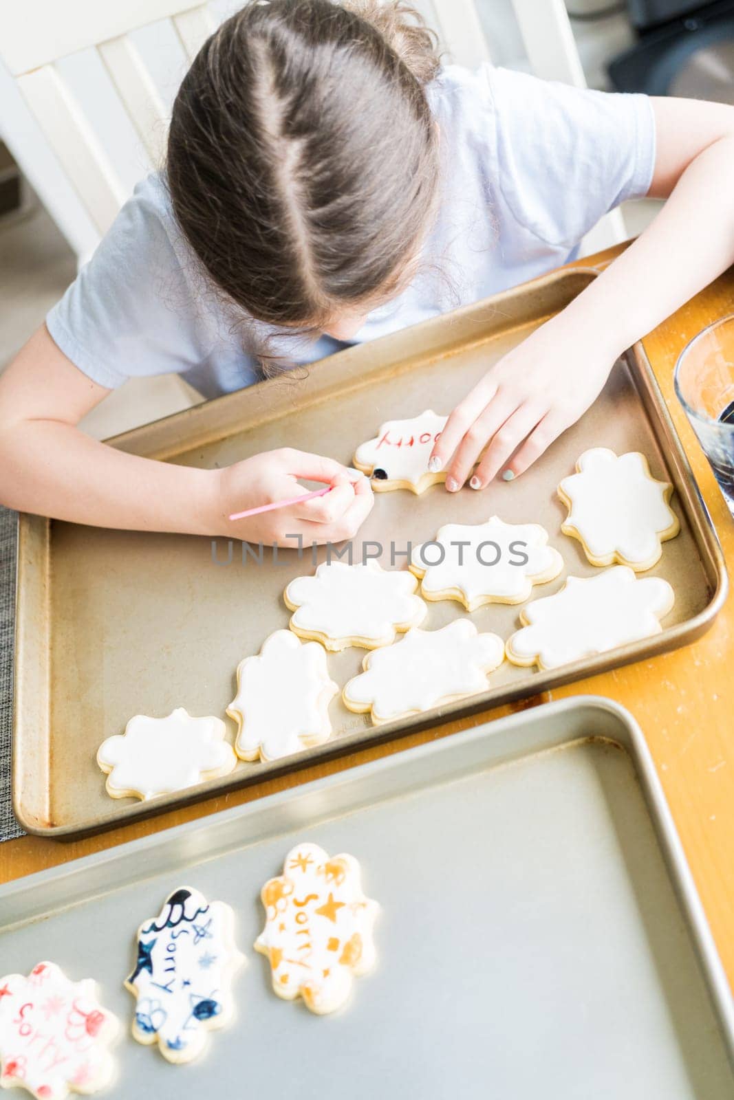 A heartwarming scene of a little girl carefully writing 'Sorry' on sugar cookies with food coloring, the cookies beautifully flooded with white royal icing.