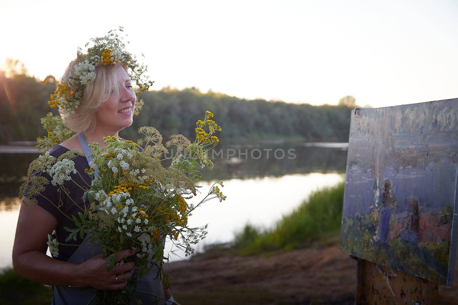 Adult female artist painting picture on the coast of water of a river or lake in nature and trees and greenery in the background