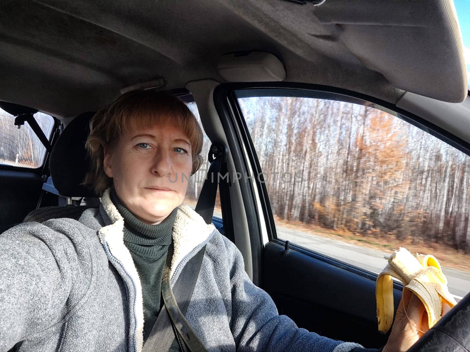 Portrait of female driver in solo journey. Adult mature middle aged woman holding steering wheel and banana. Eating while driving for to stay awake. Lady girl with food while travel or trip by car by keleny