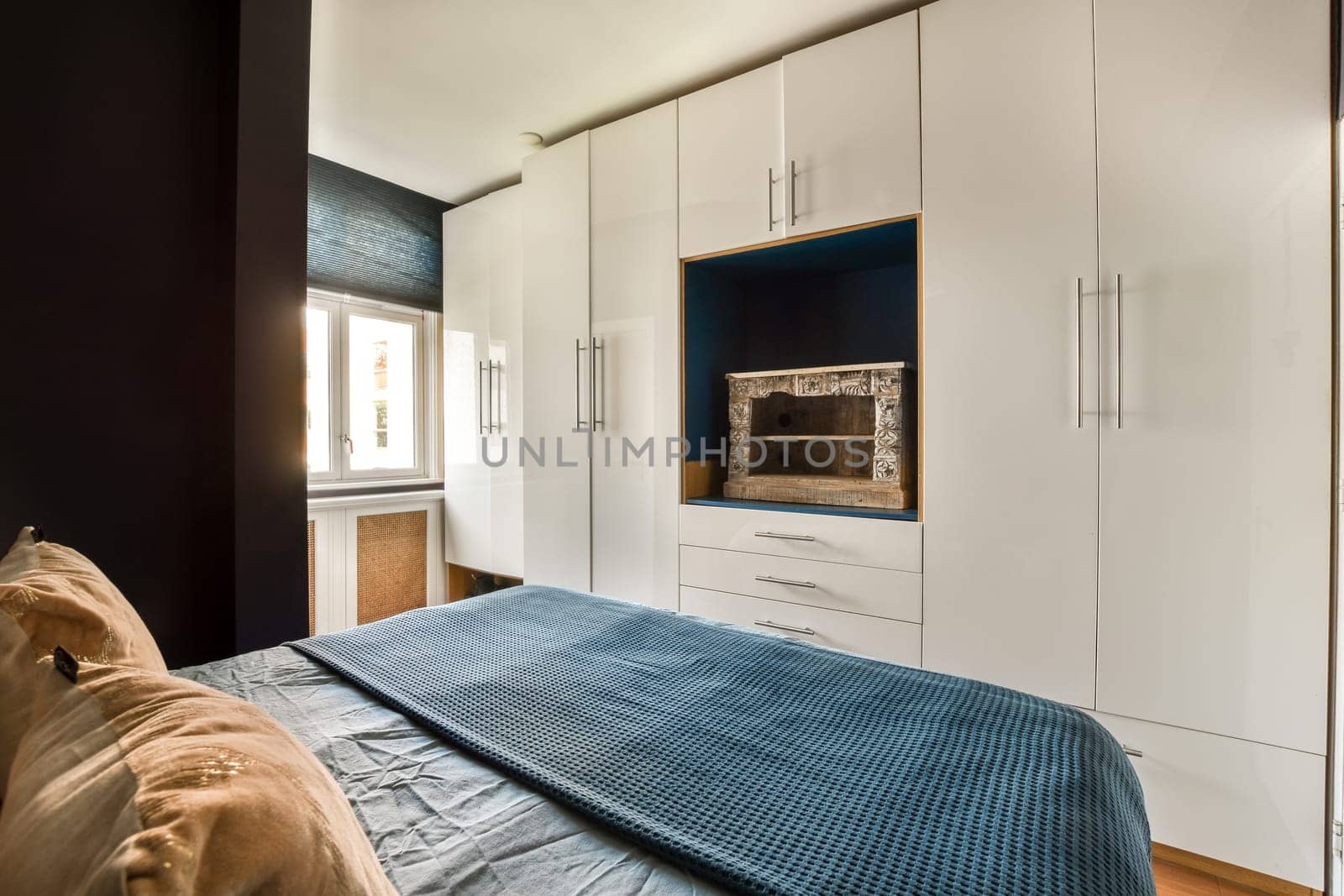 a bedroom with white cupboards and blue blanket on the bed, in front of a wall mounted flat screen