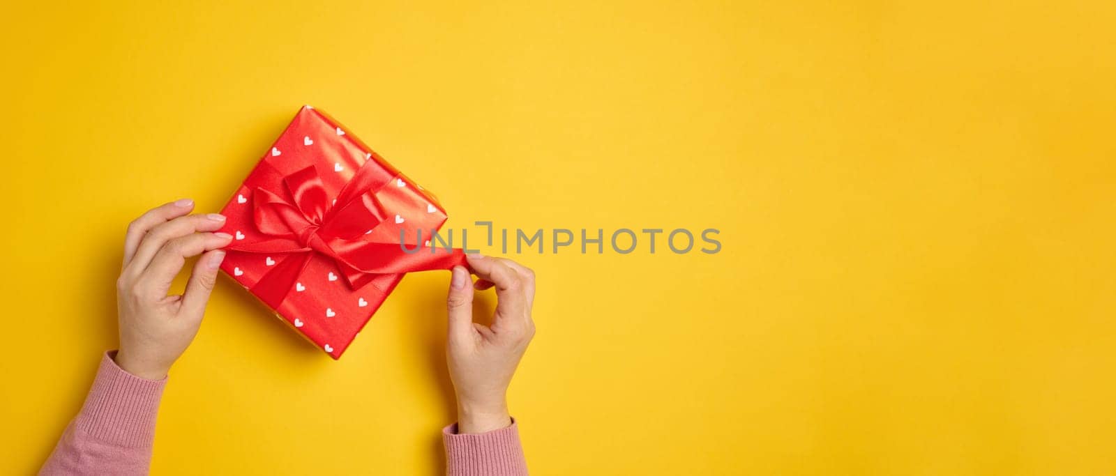 Two female hands hold a red box tied with a ribbon on a yellow background, top view. by ndanko