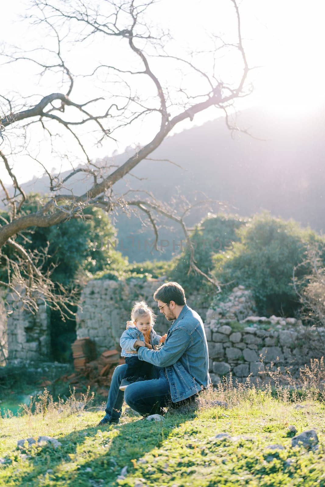 Dad with a little girl in his arms sits on a stone on a green lawn near a stone fence. High quality photo