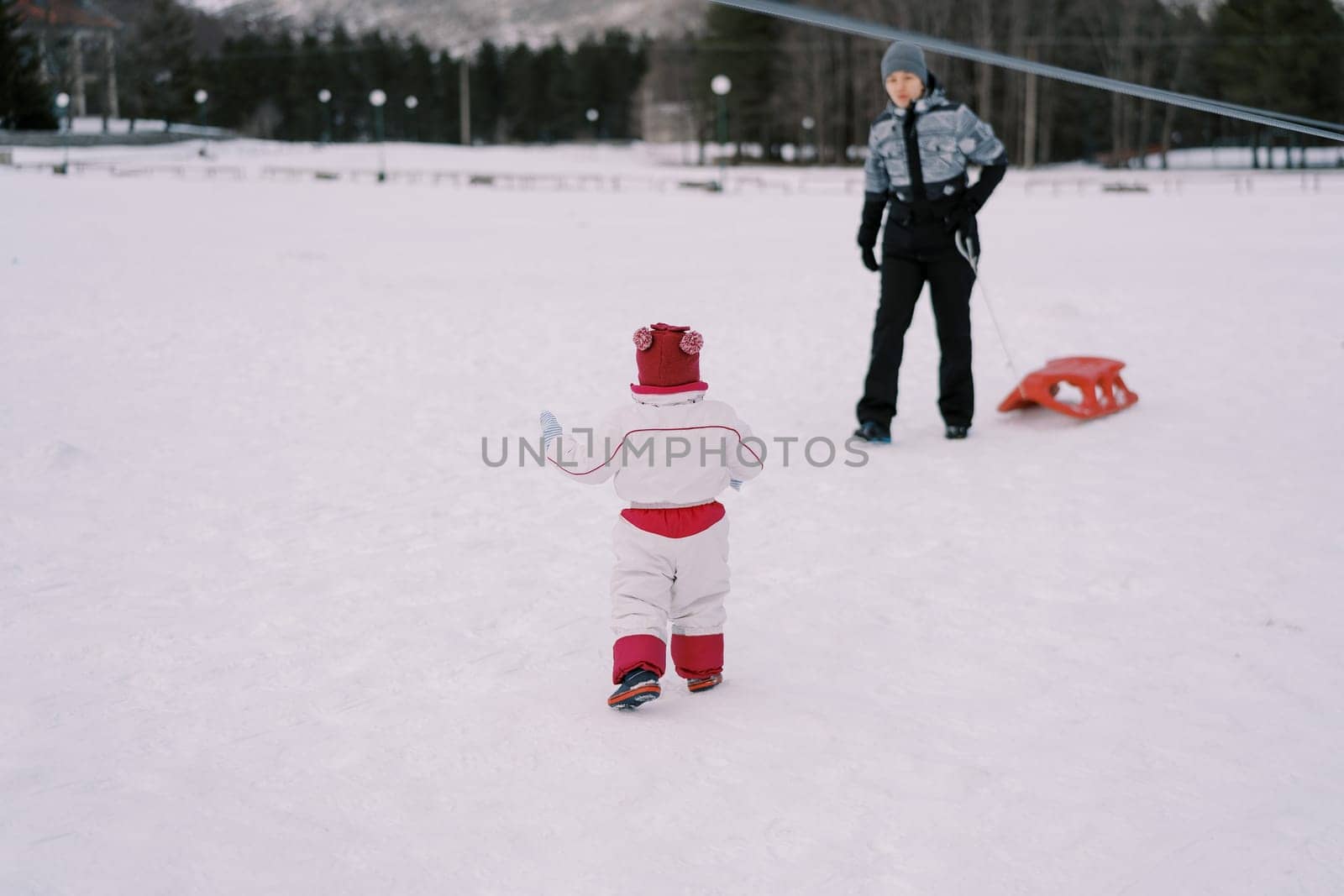 Little girl goes to her mother standing with a sled in the snow. Back view. High quality photo