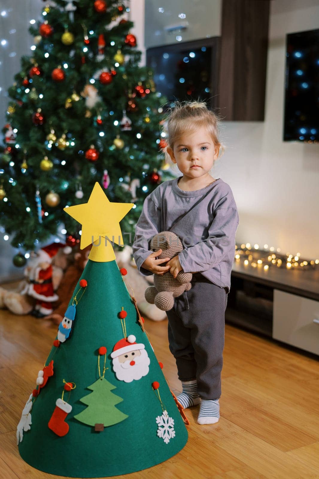 Little girl stands with a teddy bear near a decorated toy Christmas tree. High quality photo