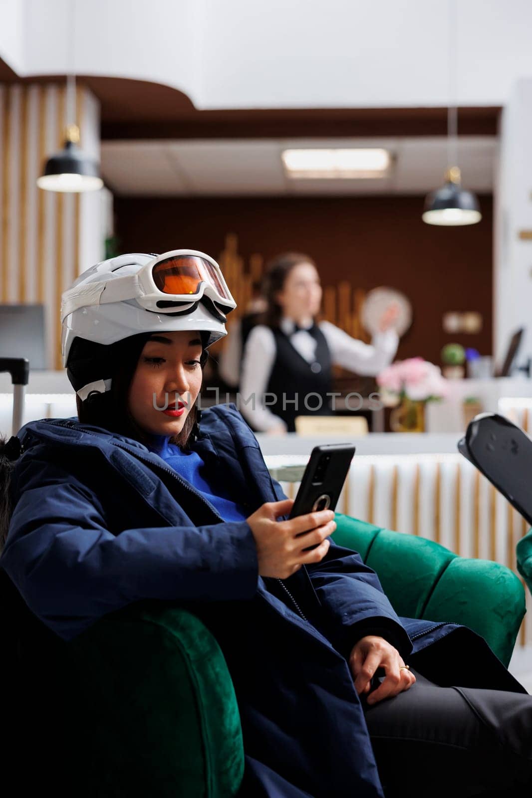 During winter holiday young asian woman uses mobile phone in cozy lounge area. Female guest sitting on sofa with digital smart device and skiing skis wearing winter jacket ski goggles and helmet.