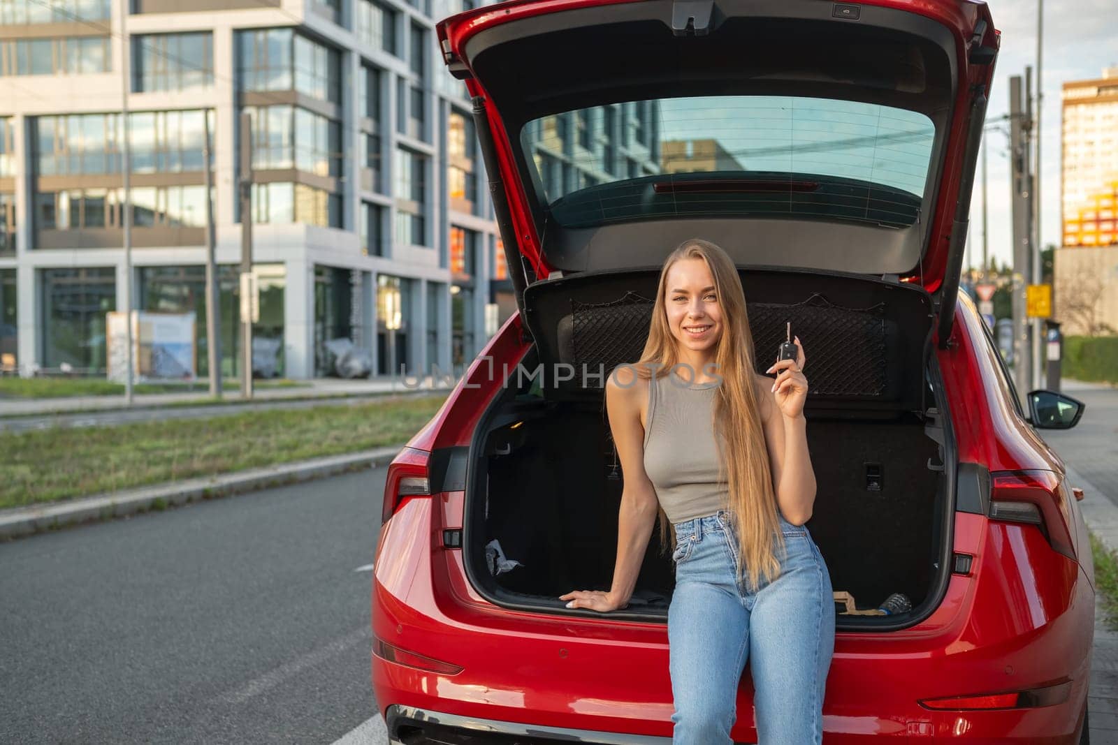 Young blonde woman sitting in the trunk of new red car and holding keys.