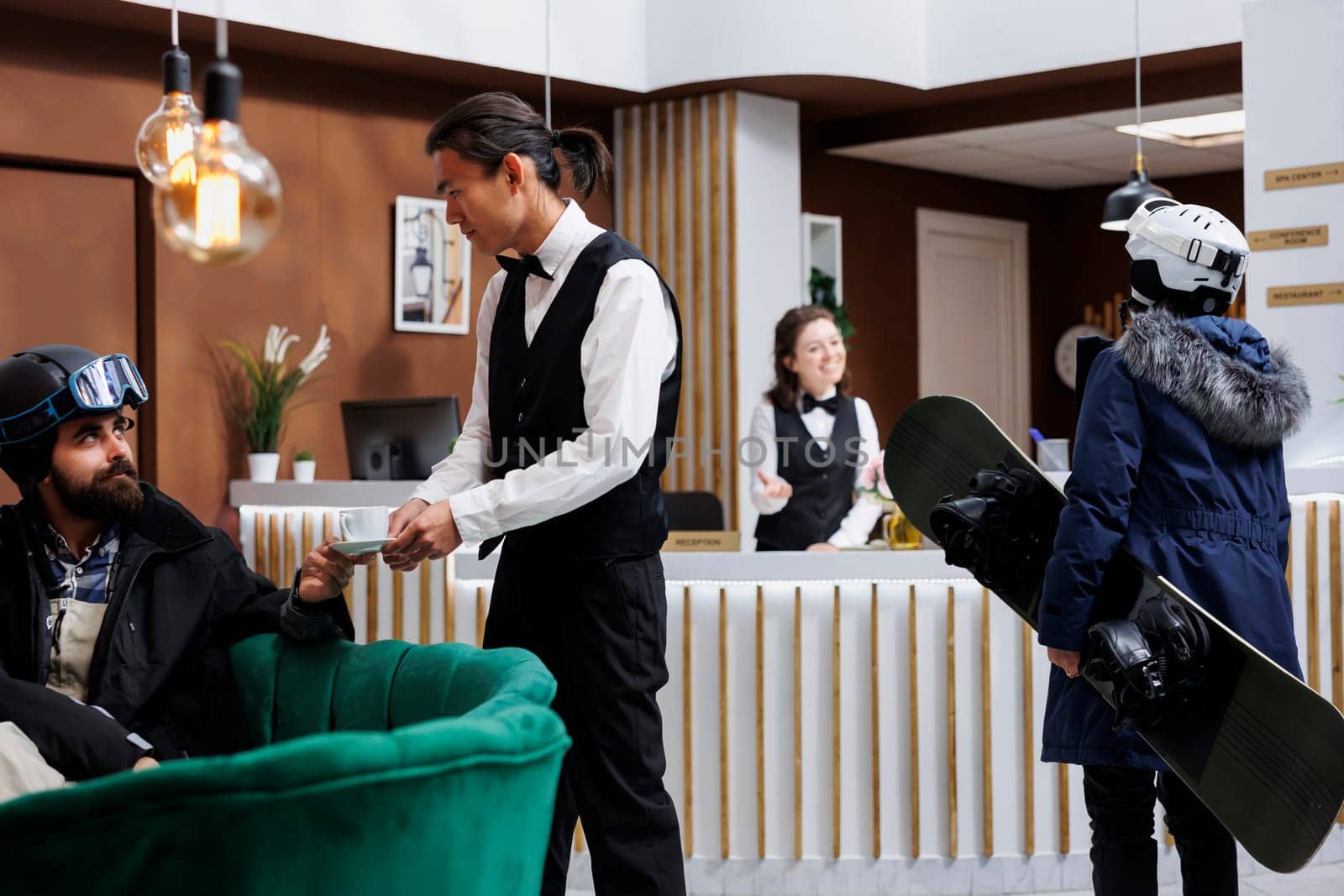 Arrival of female tourist carrying snowboard in hotel lobby for check-in with staff. Young waiter serving coffee to man on sofa. Winter holiday atmosphere with ski gear. Ski resort ensures happy stay.