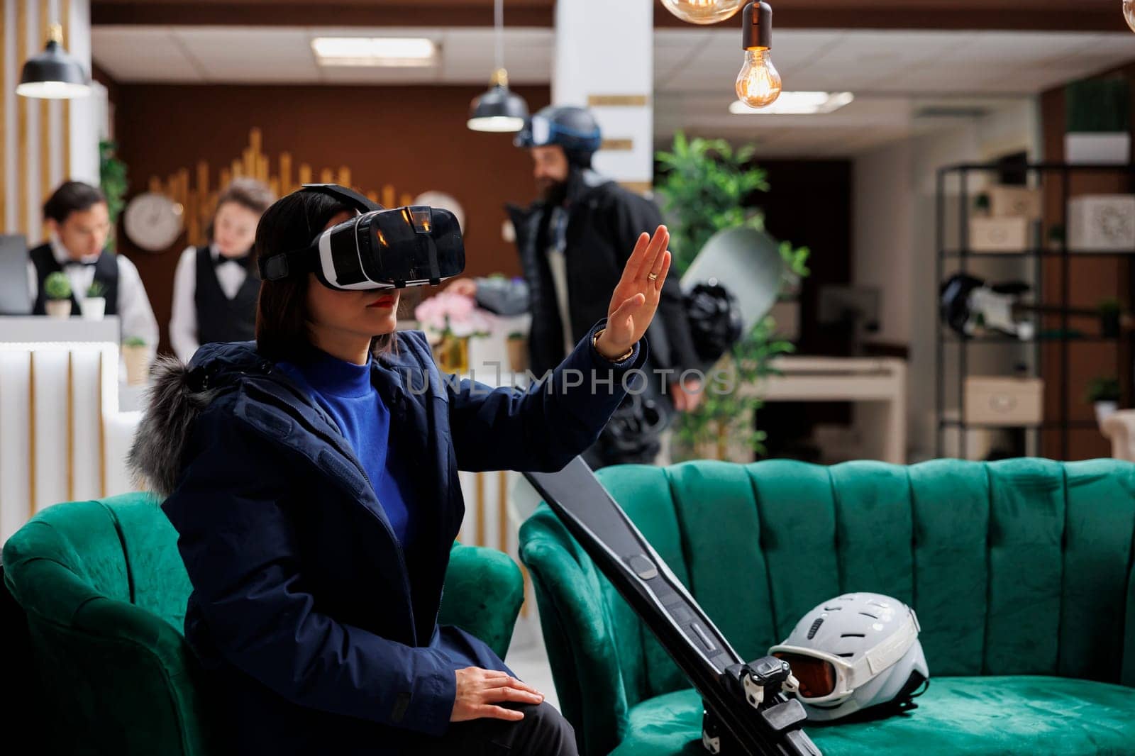 Asian woman with skiing gear engages with virtual reality device in lounge area preparing for wintersports activity. With digital VR glasses, ski resort guest balances relaxation and exploration.