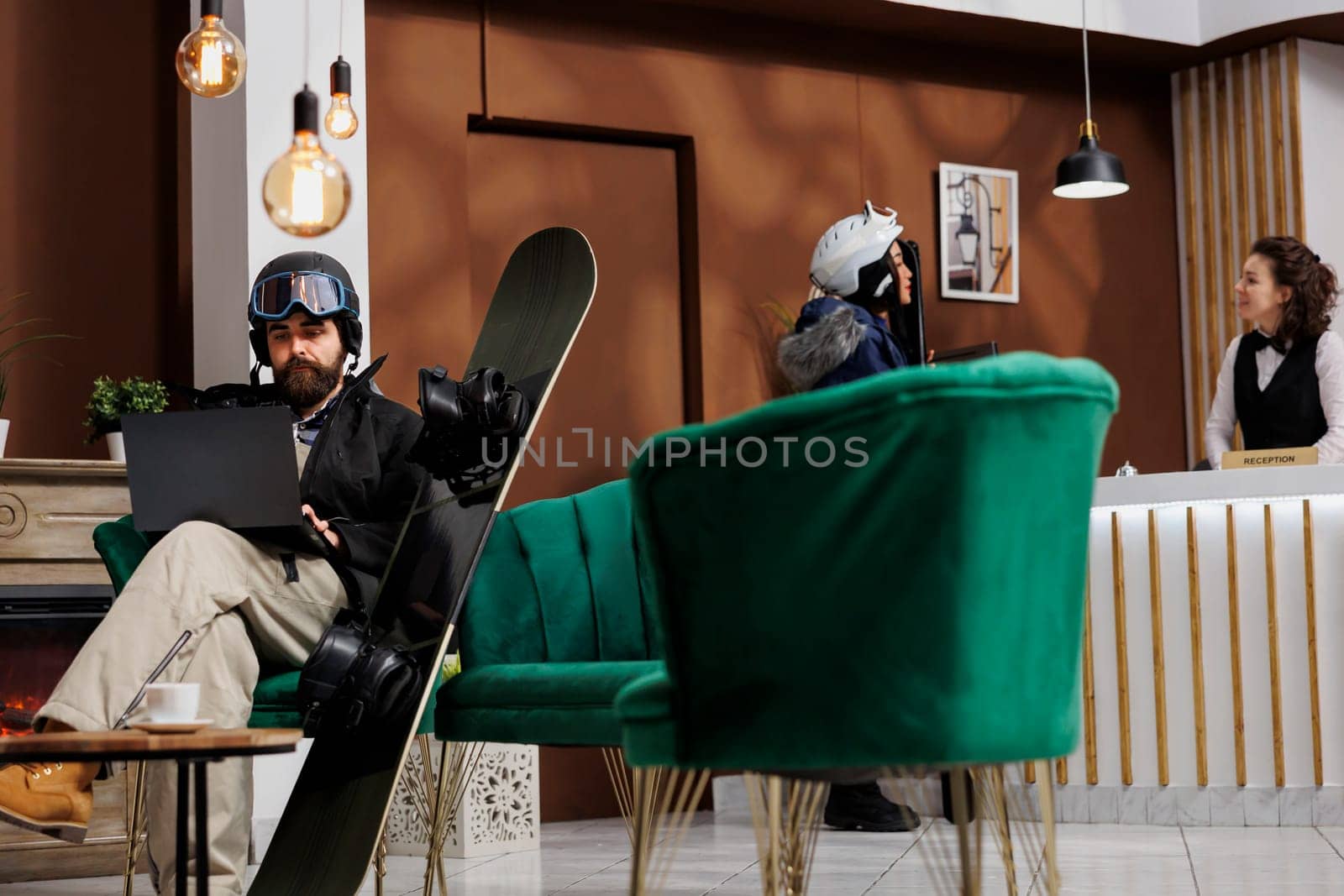 Caucasian man wearing winter clothing sitting on hotel lobby sofa and using laptop for booking. Staff assists guest in the background. Snowboard and ski goggles suggests ski resort holiday.