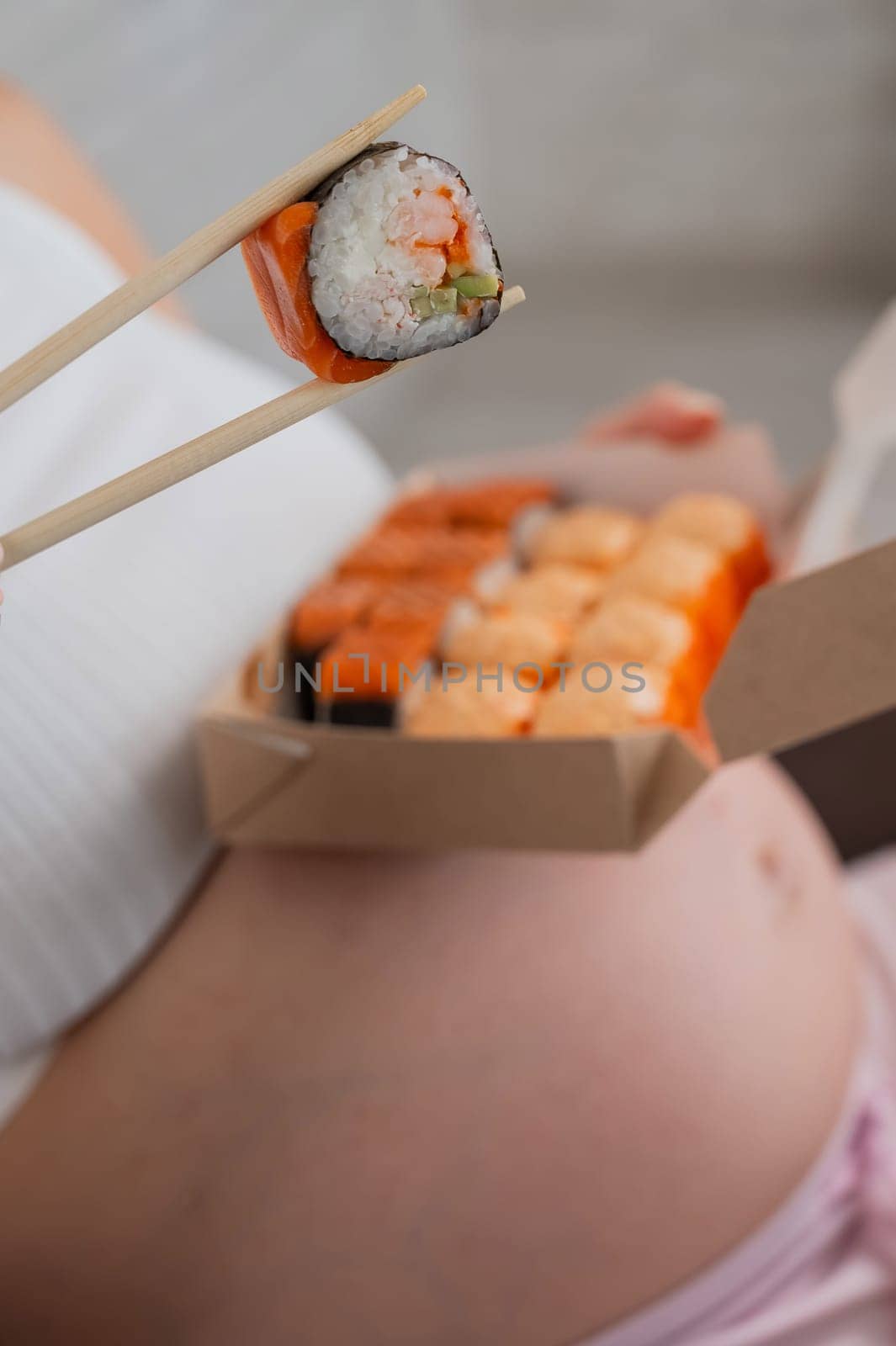 A pregnant woman sits on the sofa and eats rolls from a box. Food delivery. Close up of belly. by mrwed54