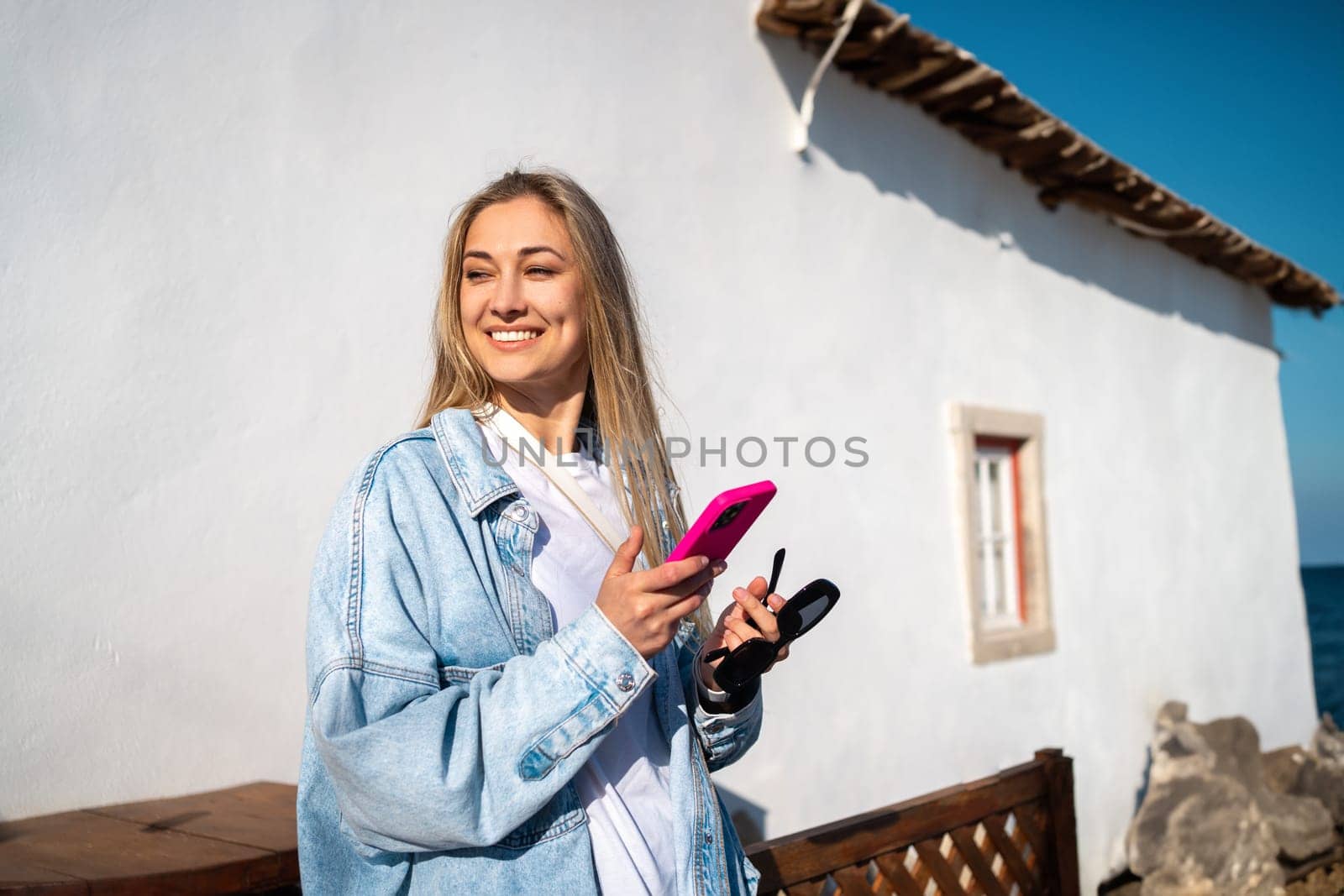 Positive female with phone and glasses outside beachfront hotel. Woman dressed denim shirt stands outside her house holding cellphone looking into distance. Happy adult woman standing at exotic resort