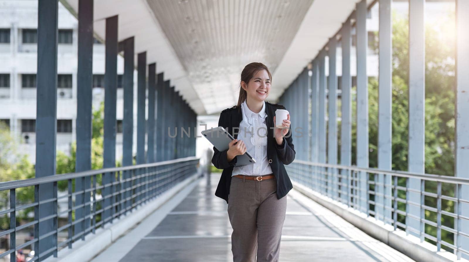 Asian businesswoman holding a laptop and a coffee mug. Smiling at the camera. Confident female employee holding a laptop standing in front of the camera on the background of a city skywalk..
