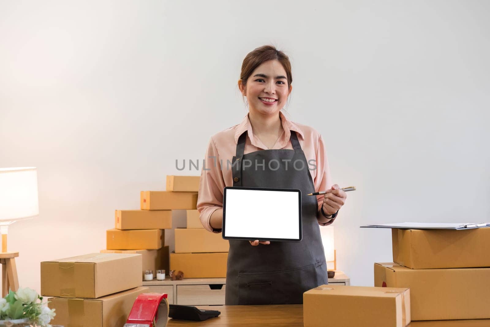 Happy Asian businesswoman holding laptop or tablet with blank screen standing in warehouse office with boxes of merchandise. Look at the camera and smile in a friendly way. by wichayada
