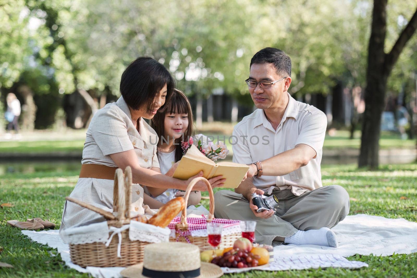 Family of senior couple and daughter picnicking in the park showing love Or reconnect after retirement in a relaxing park. An elderly man and a woman have fun on a mat in the backyard. by wichayada