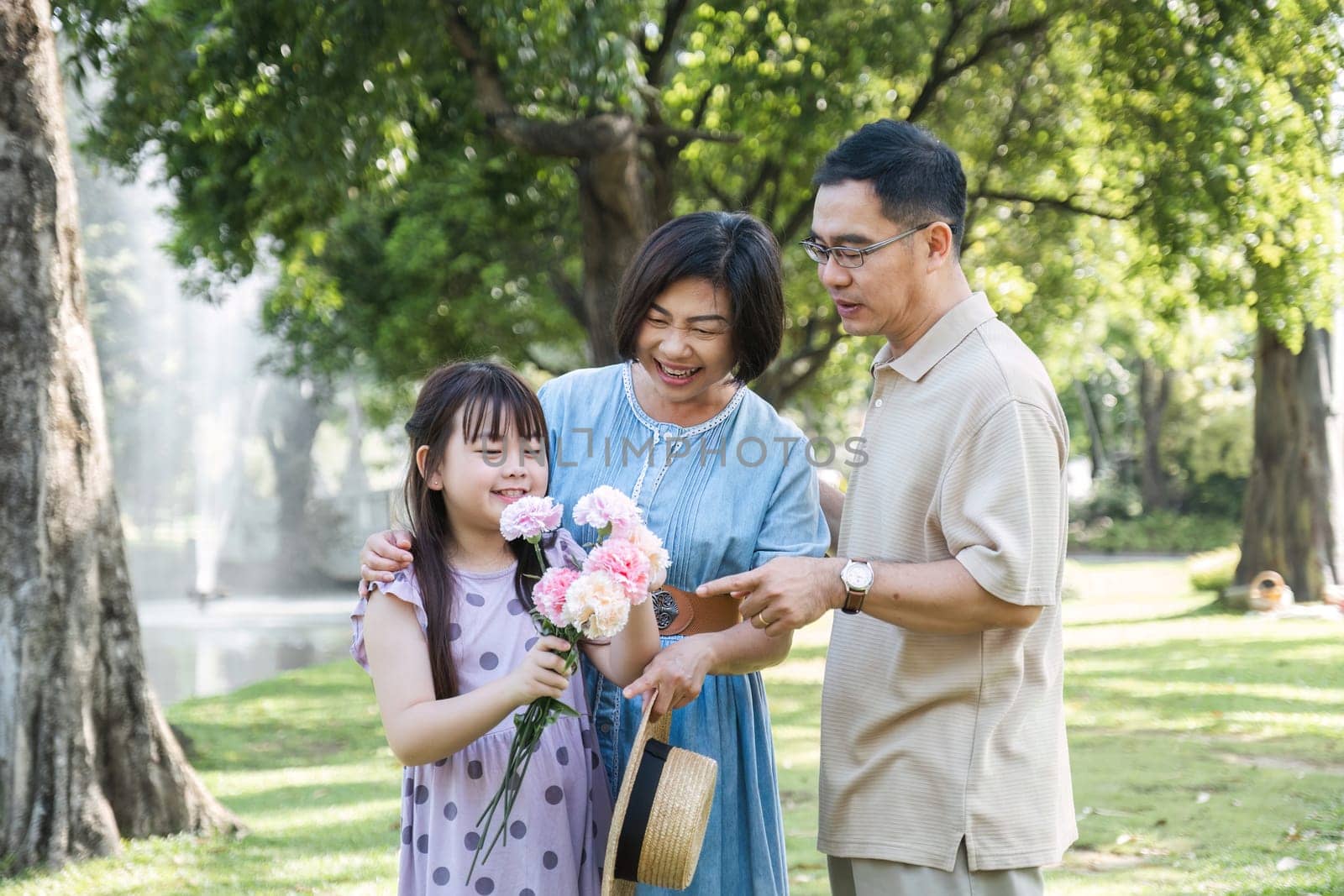 Kind-hearted Asian senior grandparent Warm and happy enjoying a stroll through the park with their adorable little granddaughter. On a clear day together lovely family by wichayada