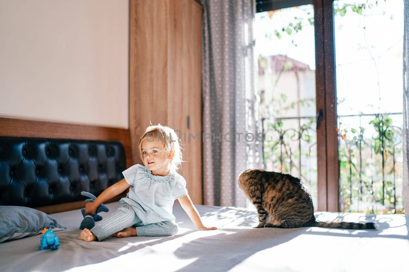 Little girl sits on a bed with a toy in her hand near a tabby cat by Nadtochiy