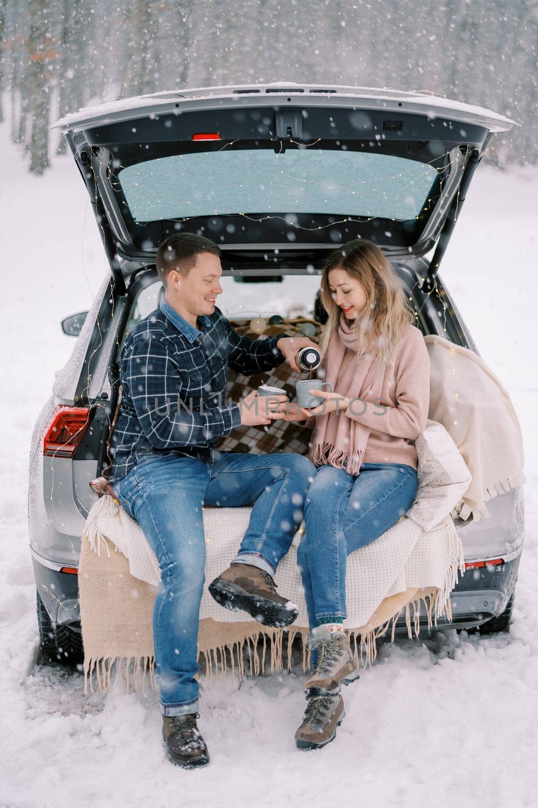 Husband pours coffee from a thermos into his wife mug while sitting with her in the trunk of a car under a snowfall in the forest by Nadtochiy