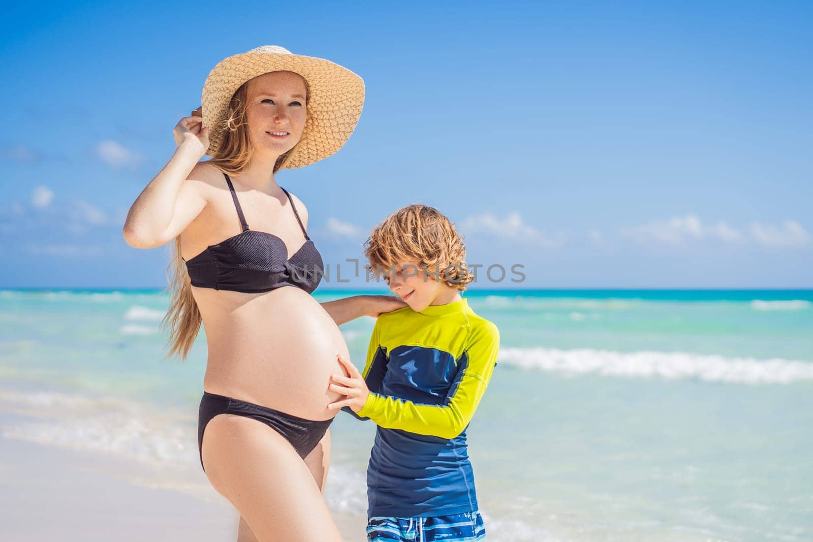 A radiant pregnant mother and her excited son share a tender moment on a serene, snow-white beach, celebrating family love amidst nature's beauty.