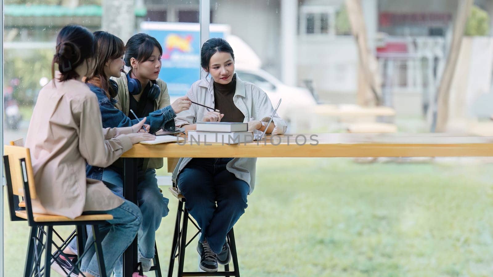Group of young Asian college students sitting on a bench in a campus relaxation area, talking, sharing ideas, doing homework or tutoring for the exam together.