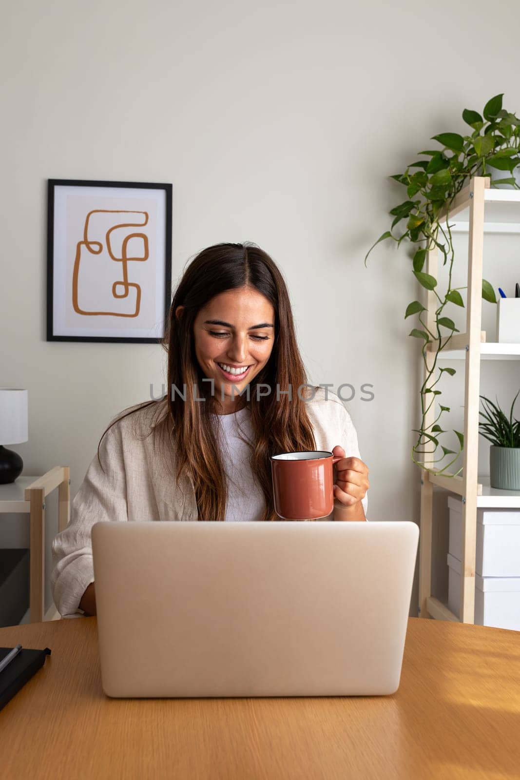 Happy young woman working at home office having coffee. Caucasian female entrepreneur using laptop. Vertical image by Hoverstock