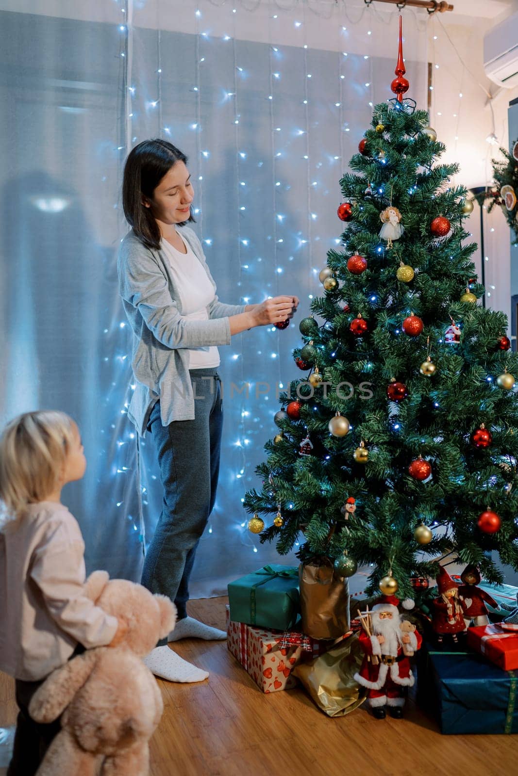 Little daughter with a teddy bear in her hand watches her mother decorate the Christmas tree. Side view. High quality photo