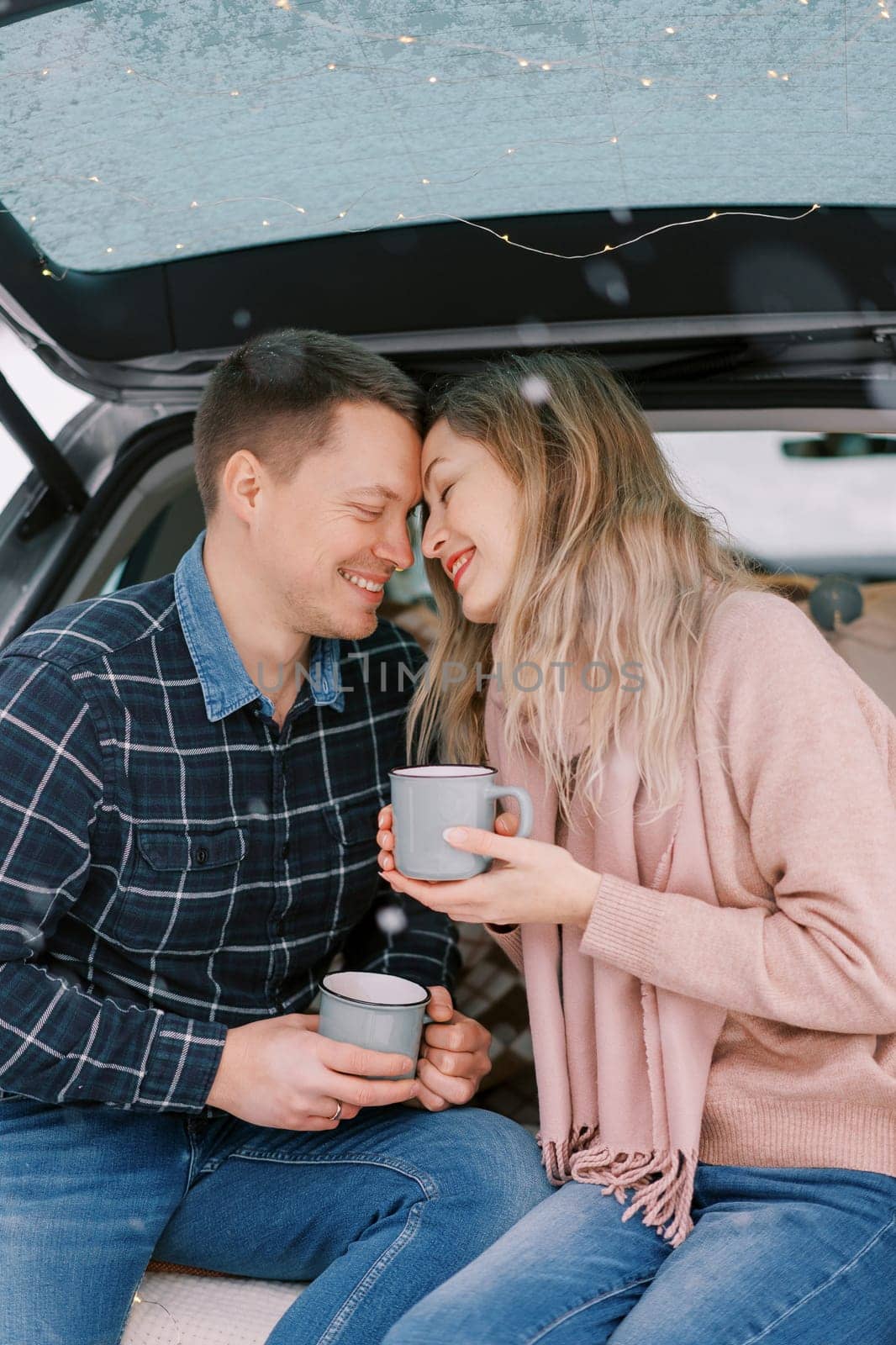 Smiling boyfriend and girlfriend touch foreheads while sitting with coffee mugs in car trunk by Nadtochiy