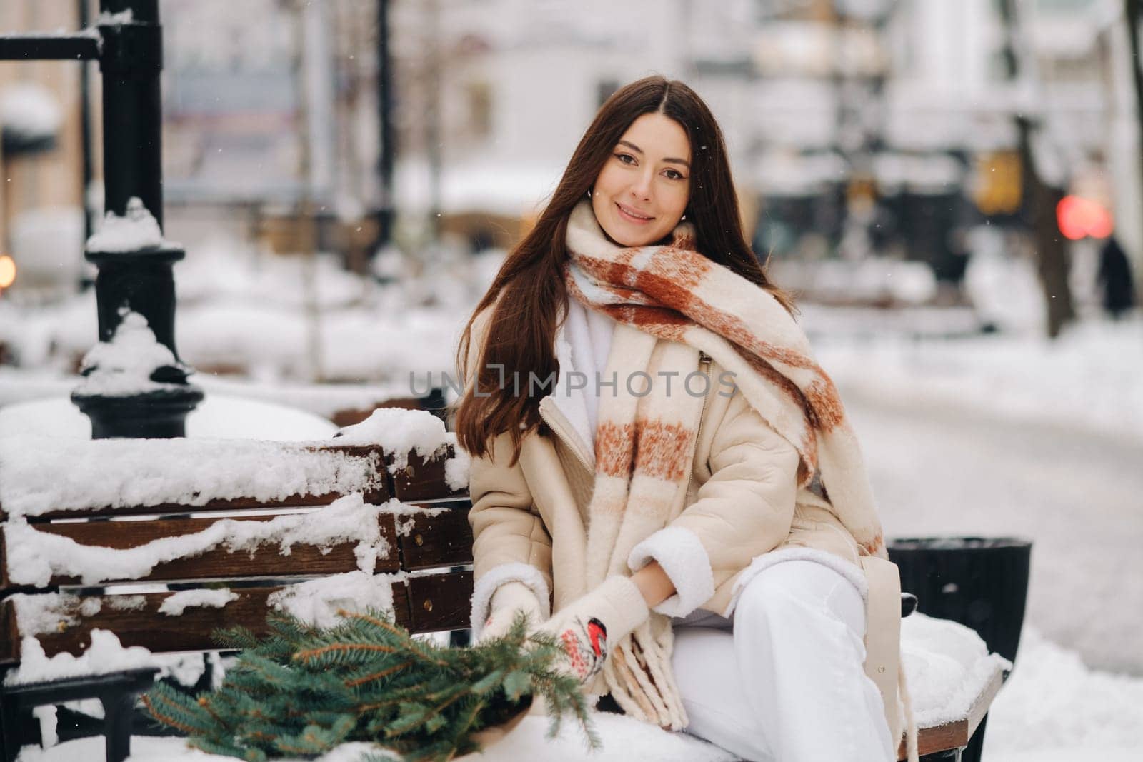 A girl with long hair in winter sits on a bench outside with a bouquet of fresh fir branches by Lobachad