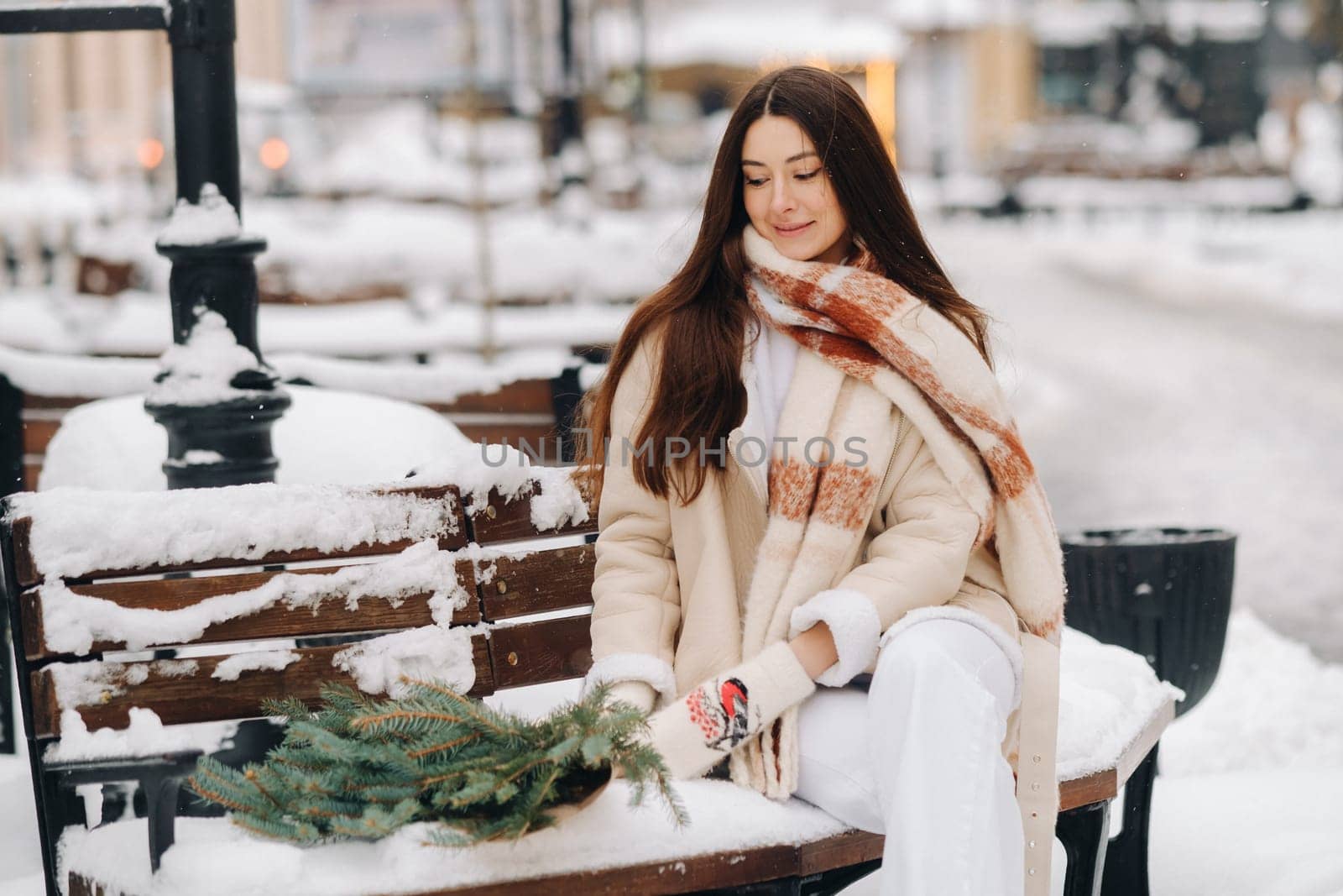 A girl with long hair in winter sits on a bench outside with a bouquet of fresh fir branches by Lobachad