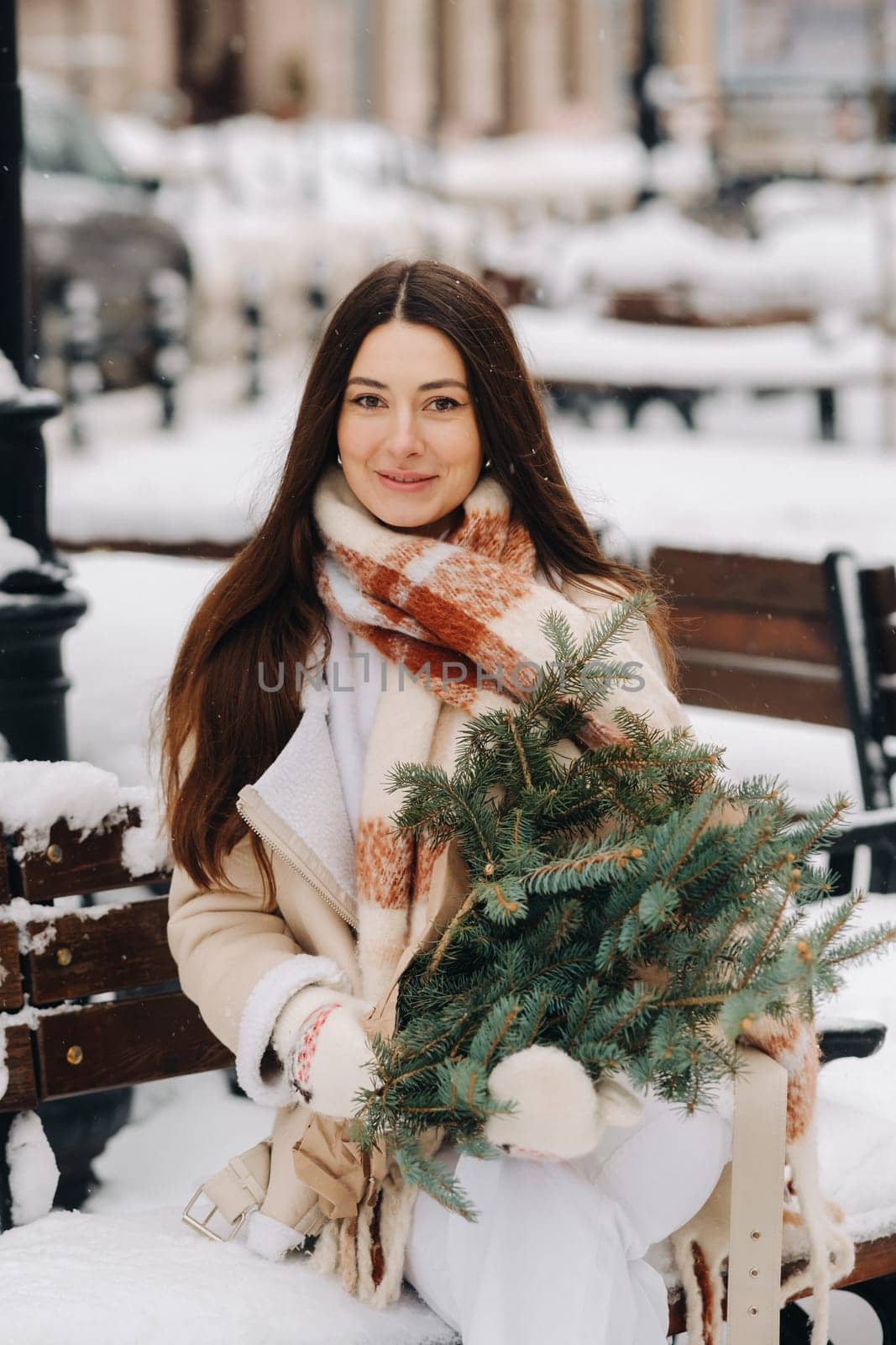 A girl with long hair in winter sits on a bench outside with a bouquet of fresh fir branches.