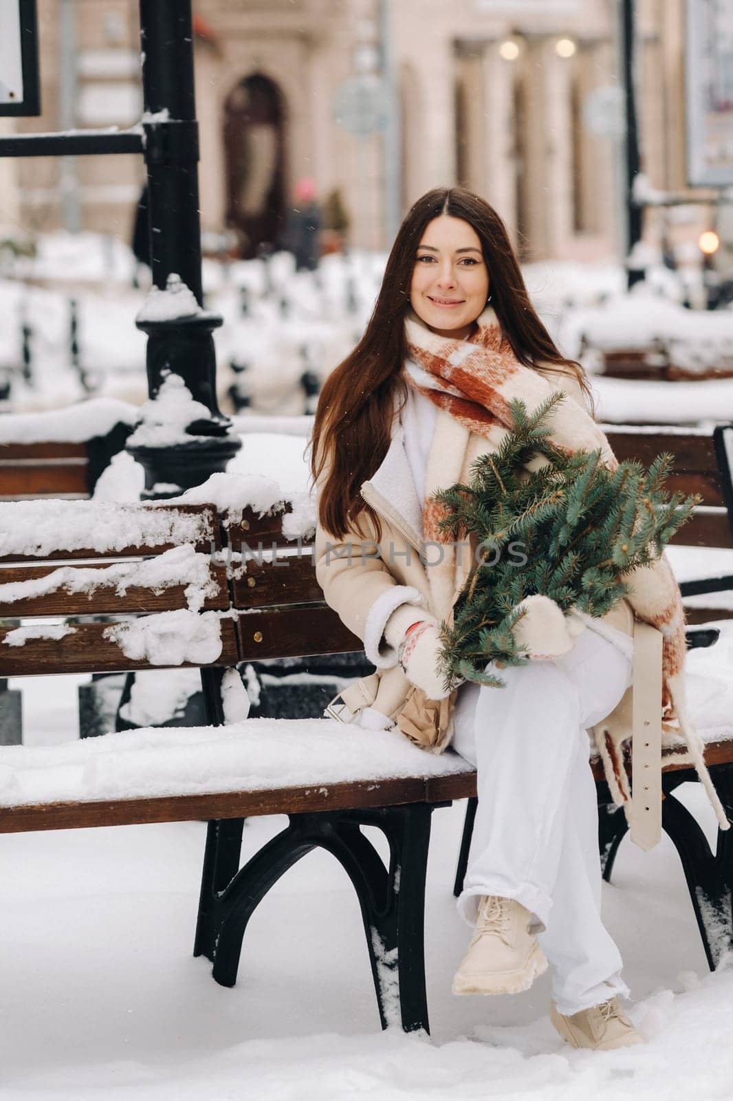A girl with long hair in winter sits on a bench outside with a bouquet of fresh fir branches.