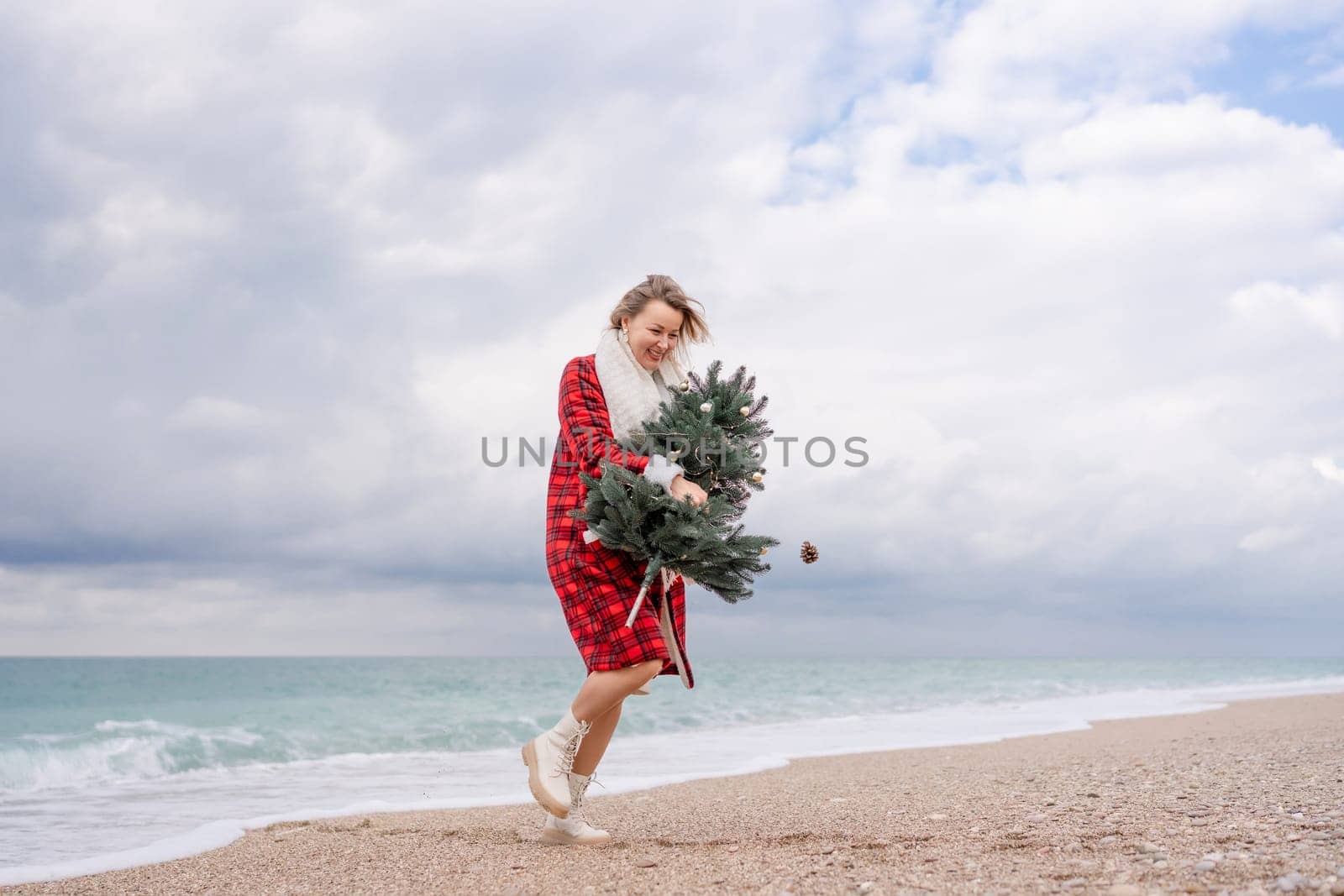 Blond woman holding Christmas tree by the sea. Christmas portrait of a happy woman walking along the beach and holding a Christmas tree in her hands. Dressed in a red coat, white dress