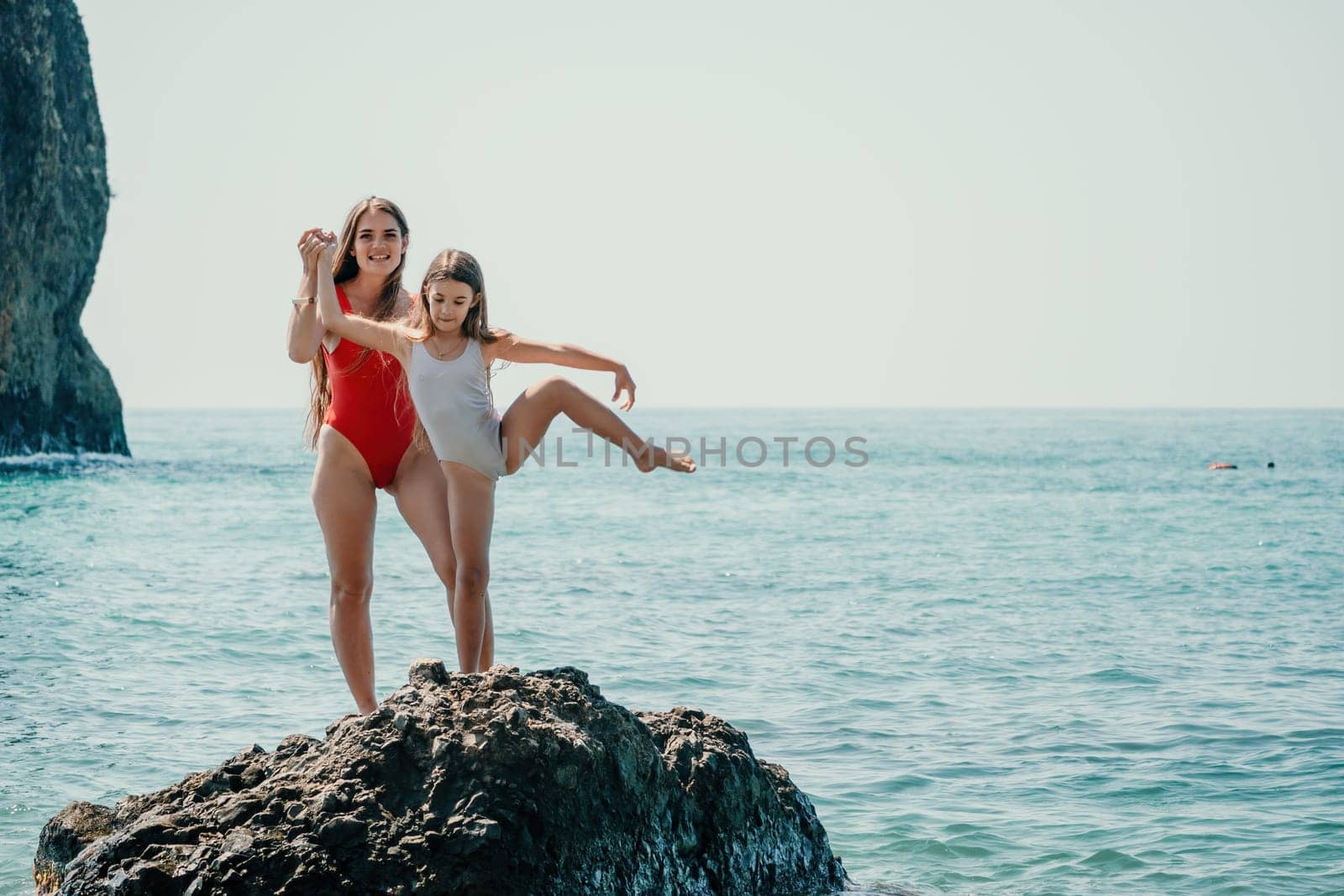 Woman and her daughter practicing balancing yoga pose on one leg up together on rock in the sea. Silhouette mother and daughter doing yoga at beach by panophotograph