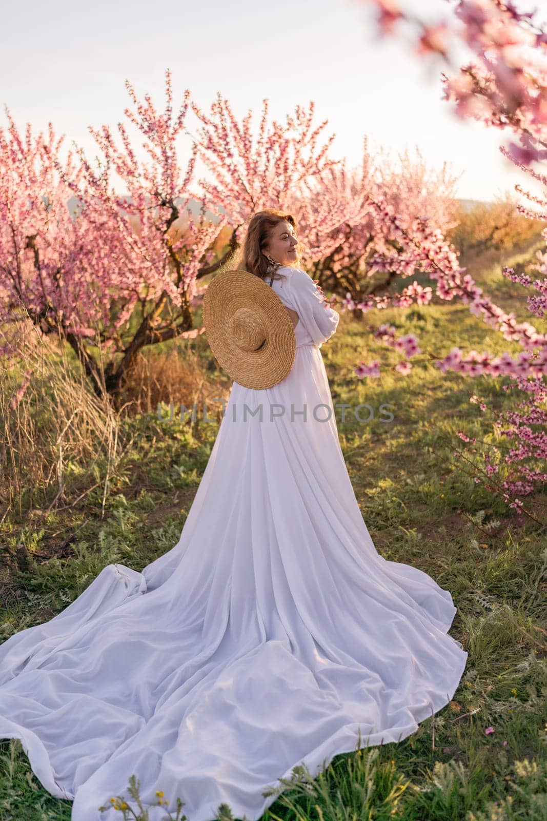 Woman blooming peach orchard. Against the backdrop of a picturesque peach orchard, a woman in a long white dress and hat enjoys a peaceful walk in the park, surrounded by the beauty of nature