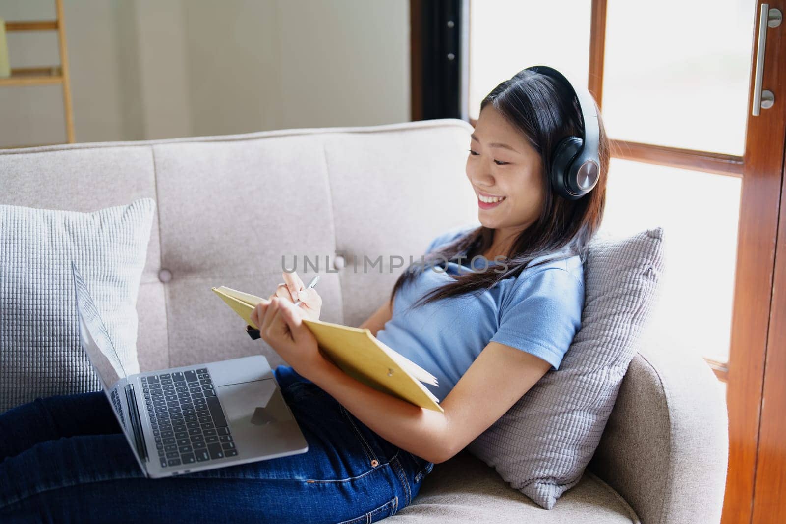 woman wearing headphones to listen music and reading notebook on sofa. by Manastrong