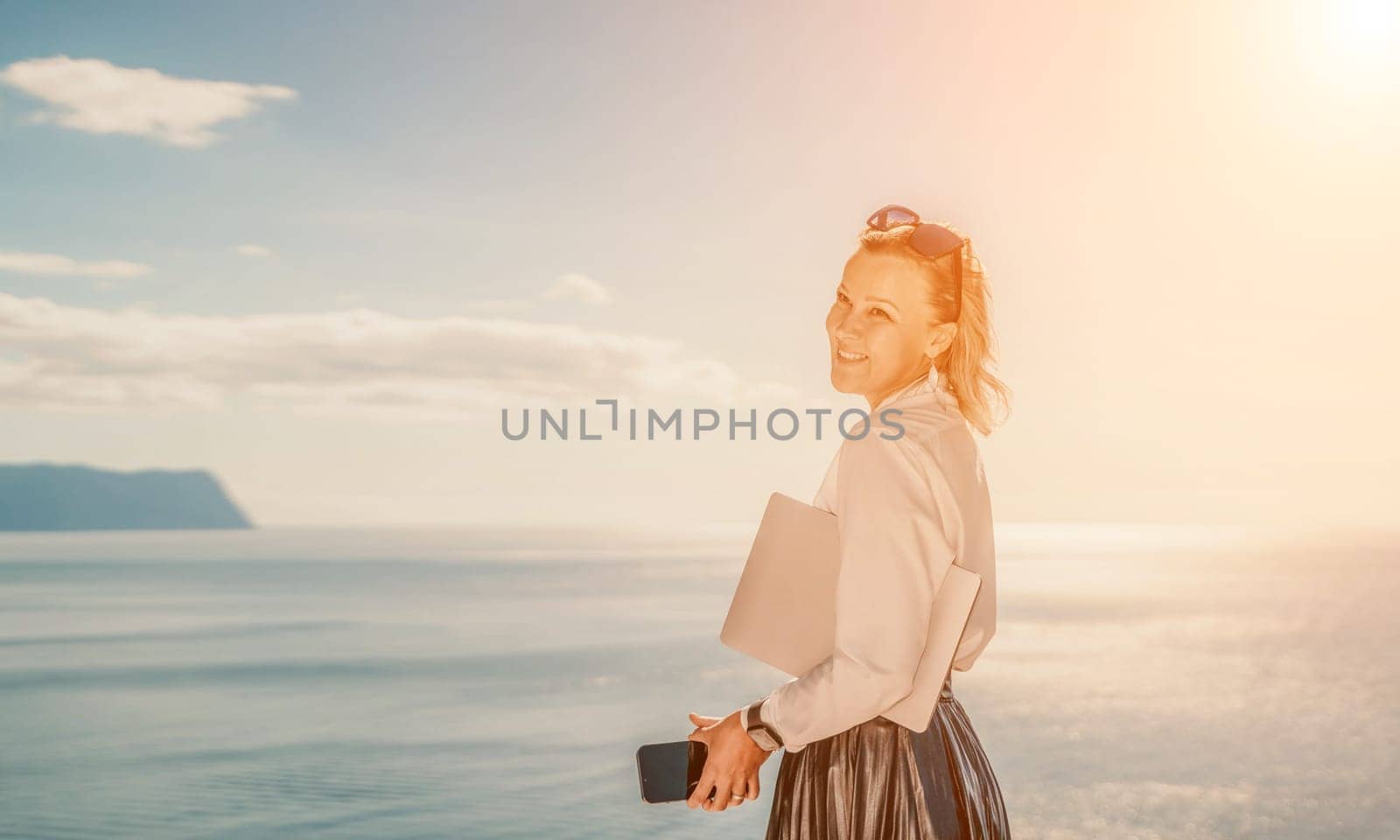 Business woman on nature in white shirt and black skirt. She works with an iPad in the open air with a beautiful view of the sea. The concept of remote work