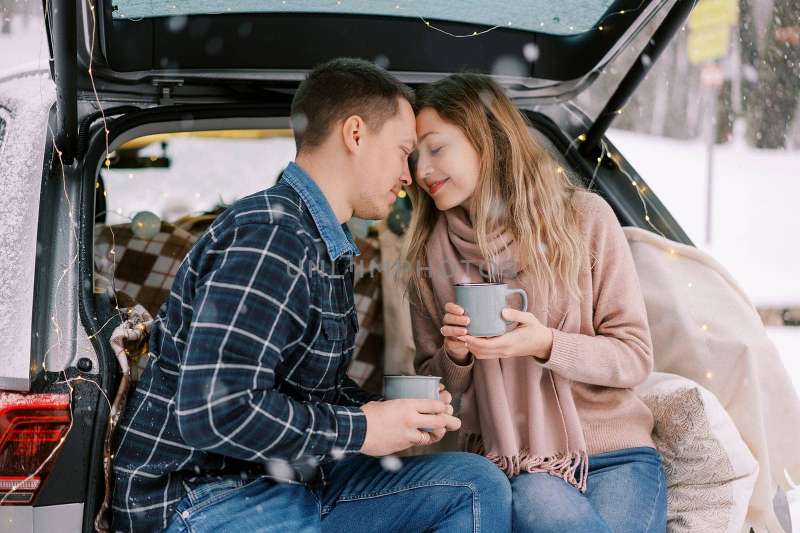 Man and woman touch foreheads while sitting with coffee mugs in car trunk in winter park. High quality photo