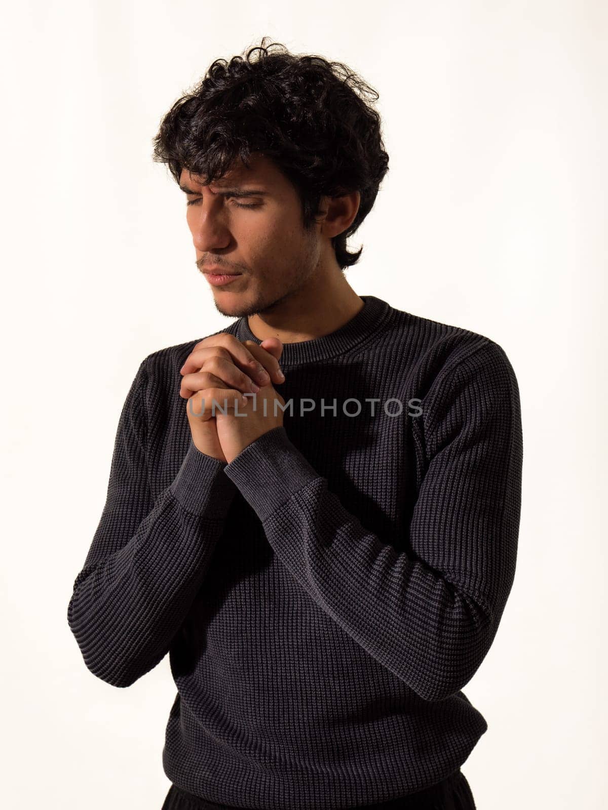 A young handsome man in a black sweater is praying, holding his hands joined together, isolated on white in studio