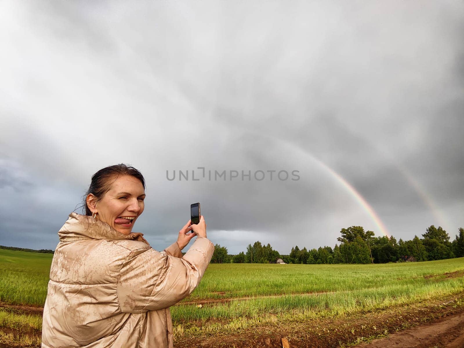 An adult girl in a field and with a stormy sky with clouds takes pictures of a rainbow and takes a selfie in the rain. A woman having fun outdoors on rural and rustic nature by keleny